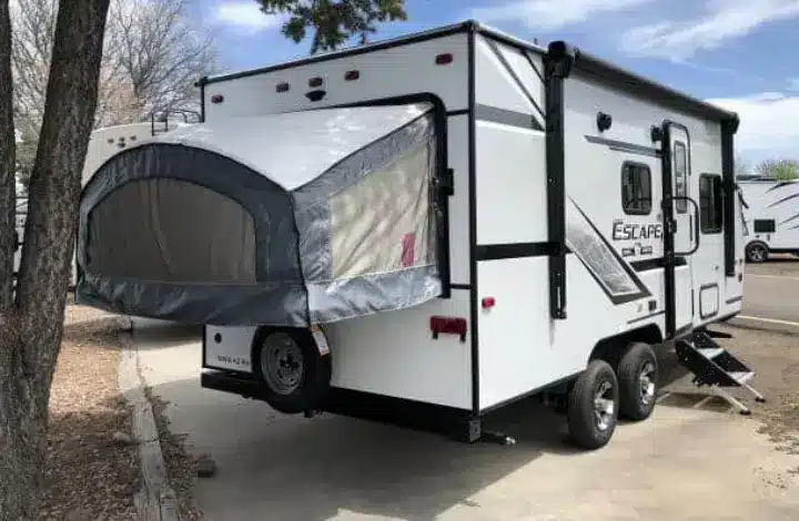 White hybrid camper trailer parked at campsite next to tree.