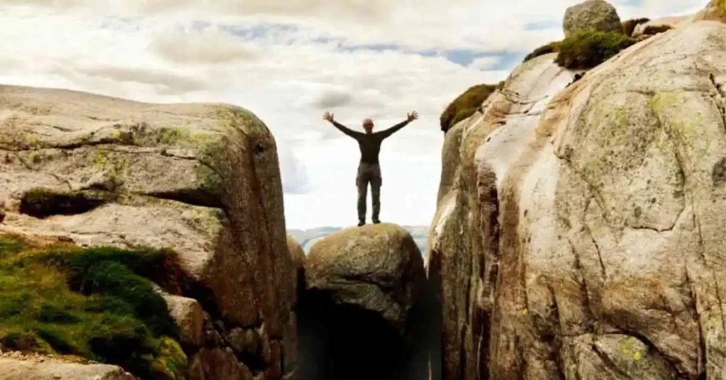 Man with arms outstretched is standing on boulder wedged between two cliffs