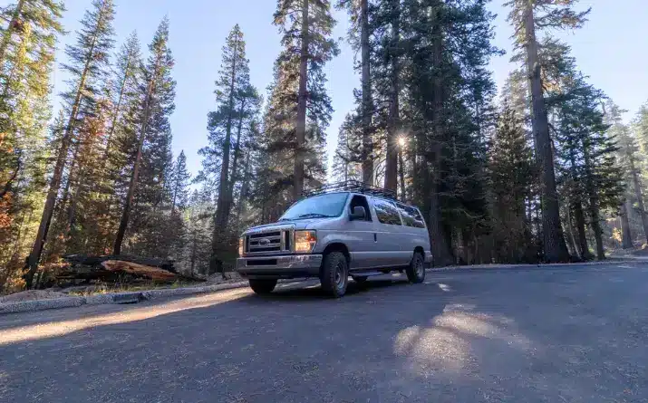 Grey campervan with lights on parked on side of road near forest.