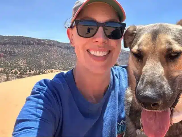 Woman wearing black sunglasses and hat sitting next to brown dog outdoors.